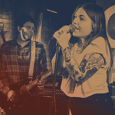 A female singing on stage in a club setting with a male guitarist in the background