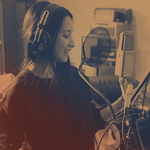 A female in an audio recording studio booth in London, Ontario with headphones on reading a script